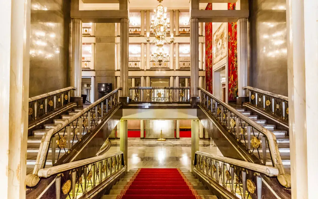 Eleganckie foyer teatru z marmurowymi schodami, złotymi balustradami i kryształowym żyrandolem. Czerwony dywan zaprasza do środka.