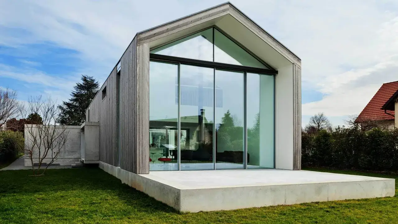 Tania nowoczesna stodoła z dużymi przeszkleniami i drewnianą elewacją, otoczona zielonym trawnikiem.