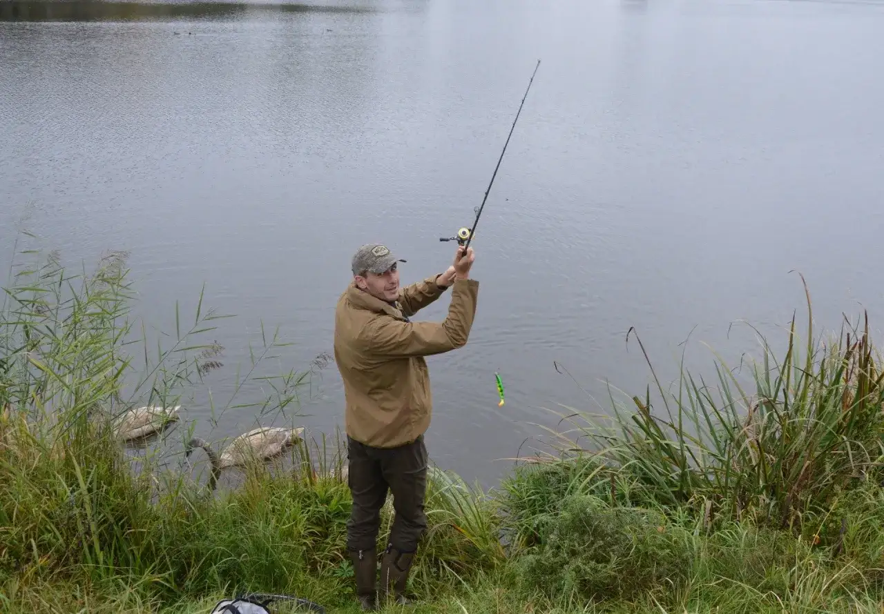 Wędkarz łowi ryby nad jeziorem, zastanawiając się, ile kosztuje karta wędkarska na całą Polskę. Obok pływają łabędzie.