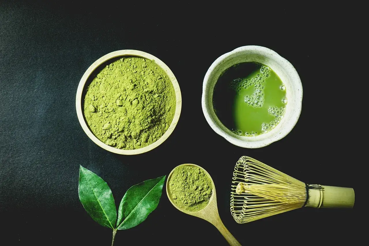 Miska z zielonym proszkiem matcha, filiżanka napoju, liście i bambusowa miotełka.