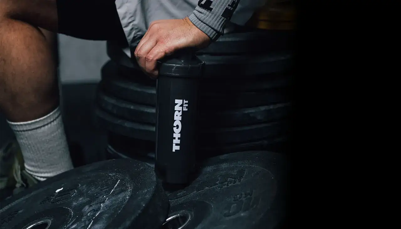 Dłoń trzyma czarny shaker z logo "THORN FIT" na tle stosu talerzy treningowych.