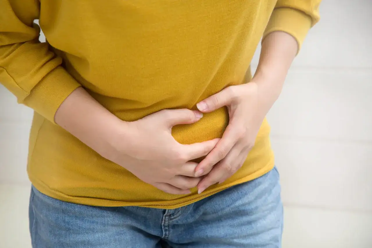 Osoba w żółtej bluzce trzyma się za brzuch, odczuwając ból. Szuka informacji, jak rozpuścić kamienie żółciowe.