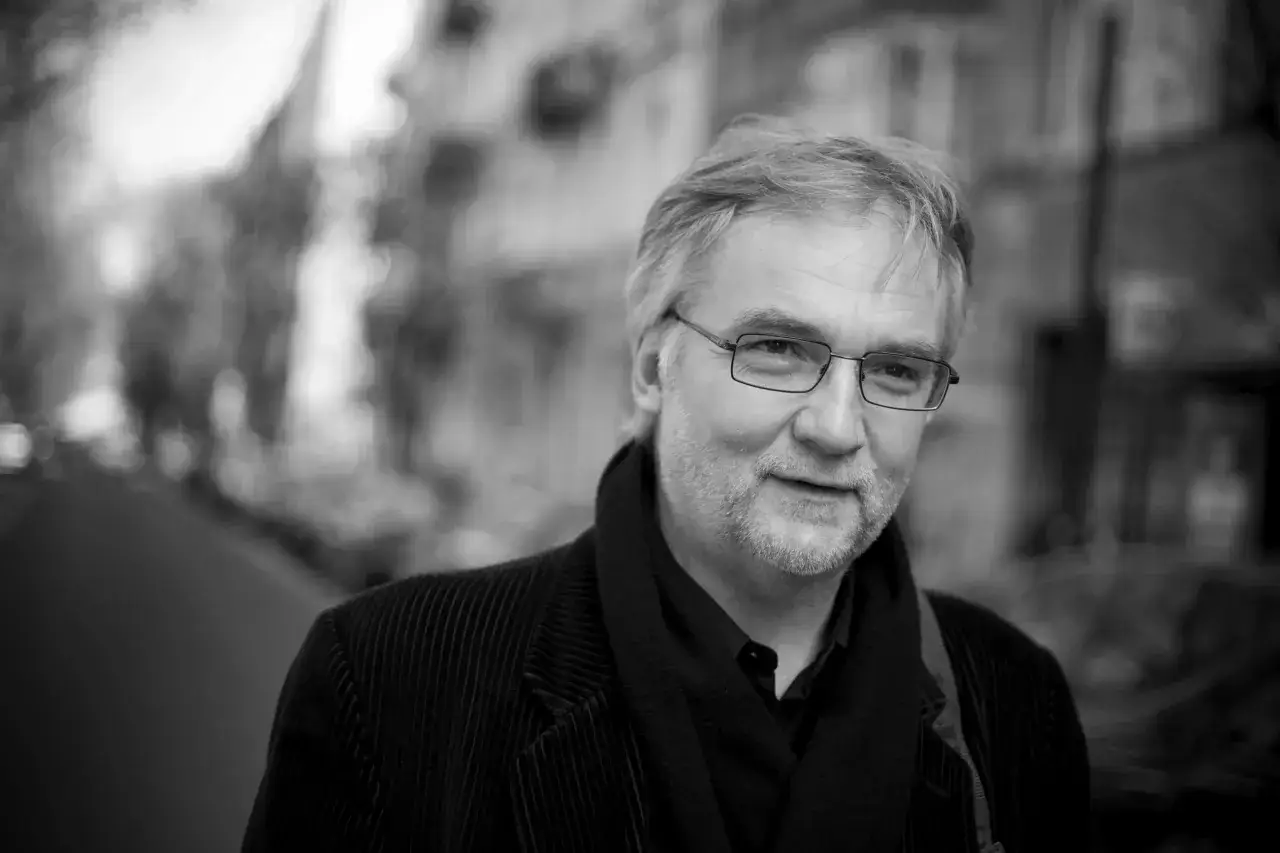 Portret Jerzego Pilcha w okularach, w czarnym sztruksowym płaszczu i szaliku.