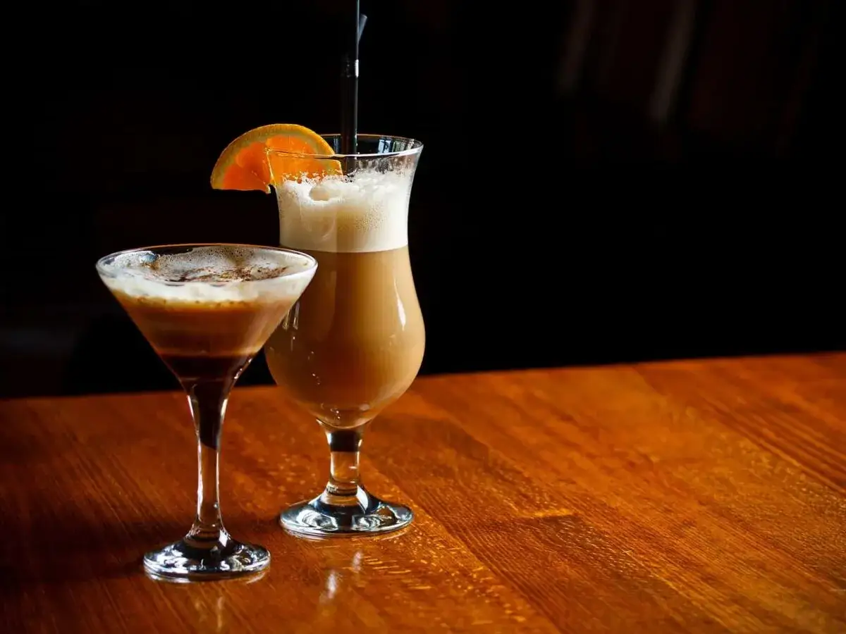 Dwa drinki z likierem kawowym i mlekiem: jeden w kieliszku martini, drugi w wysokiej szklance z pomarańczą.