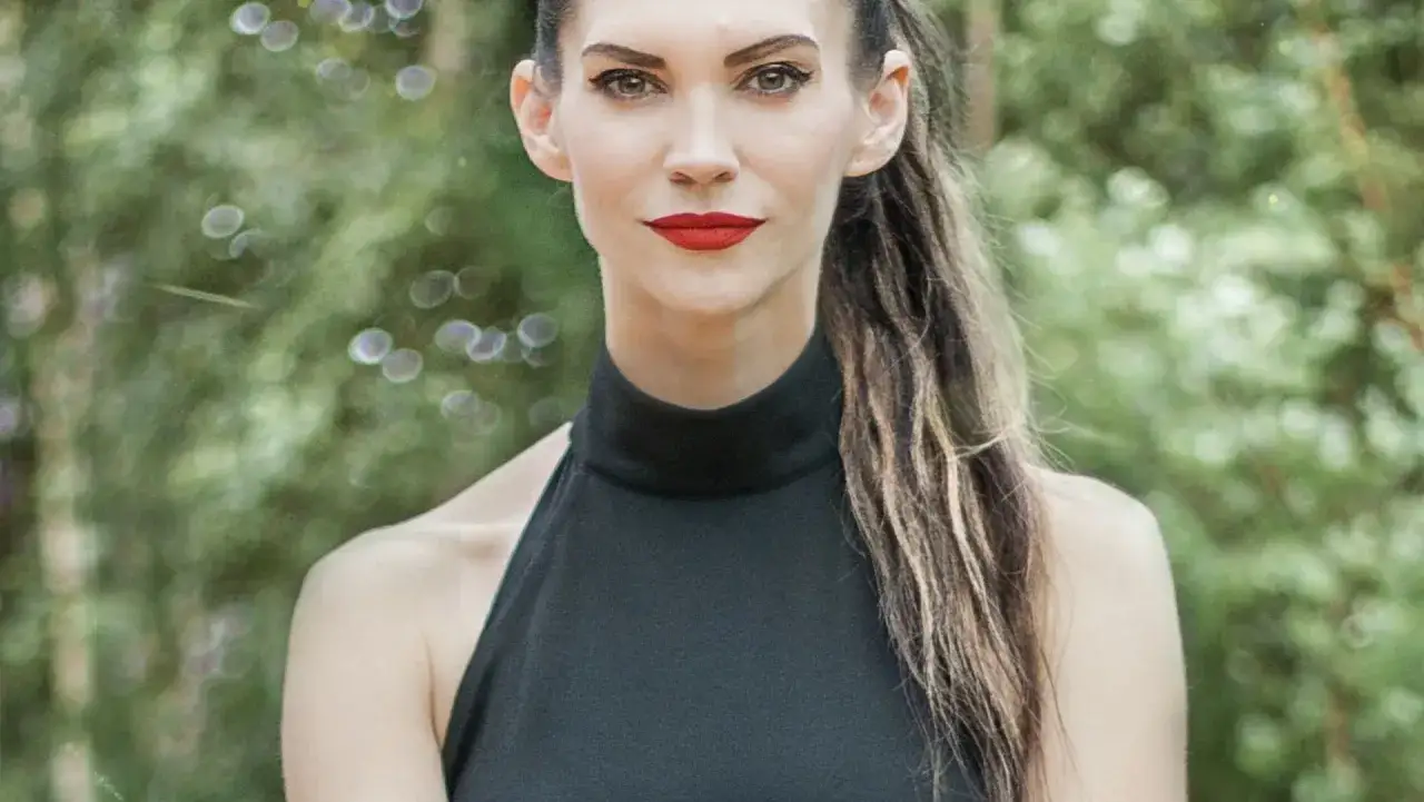 Małgorzata Oliwia Sobczak w czarnej, eleganckiej sukience, z burzą włosów i wyrazistym makijażem, na tle zieleni.
