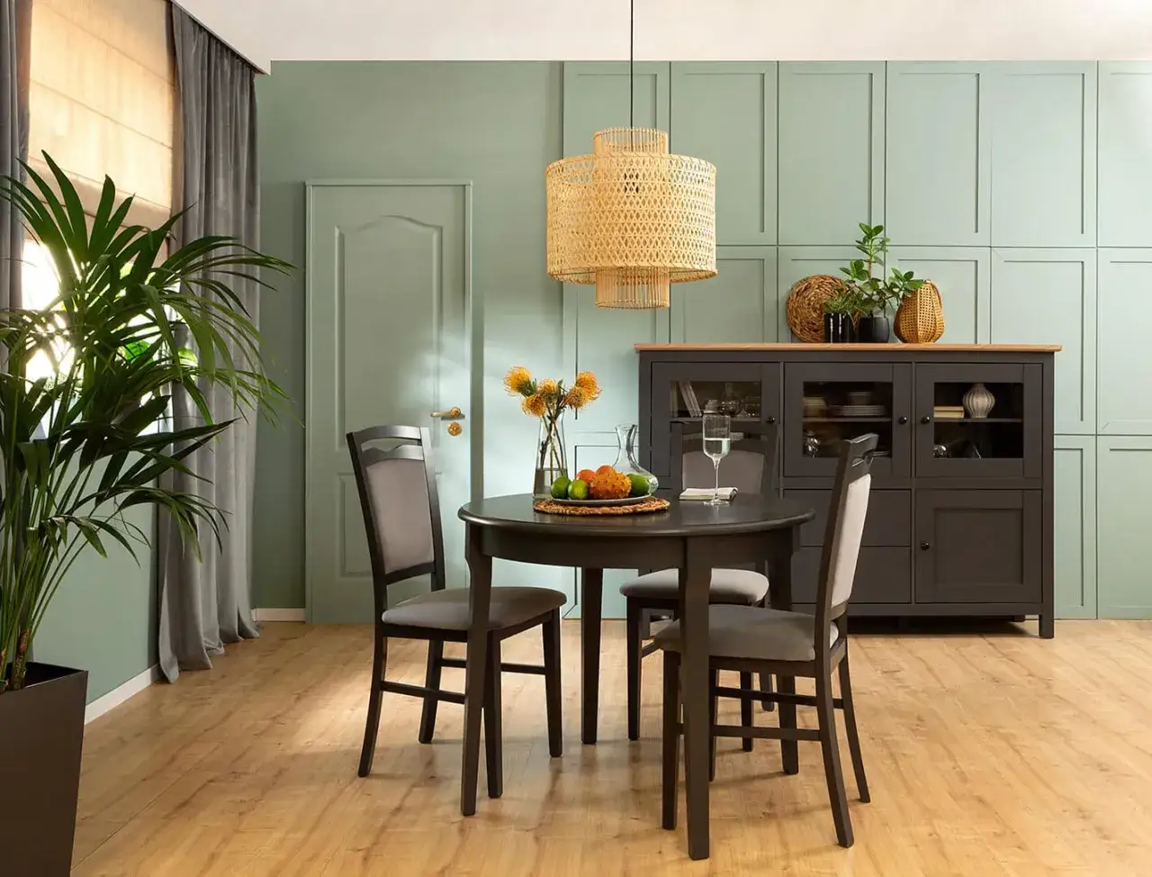 Jadalnia w odcieniu kolor szałwiowy. Okrągły stół z krzesłami, kredens i duża roślina tworzą przytulną atmosferę.