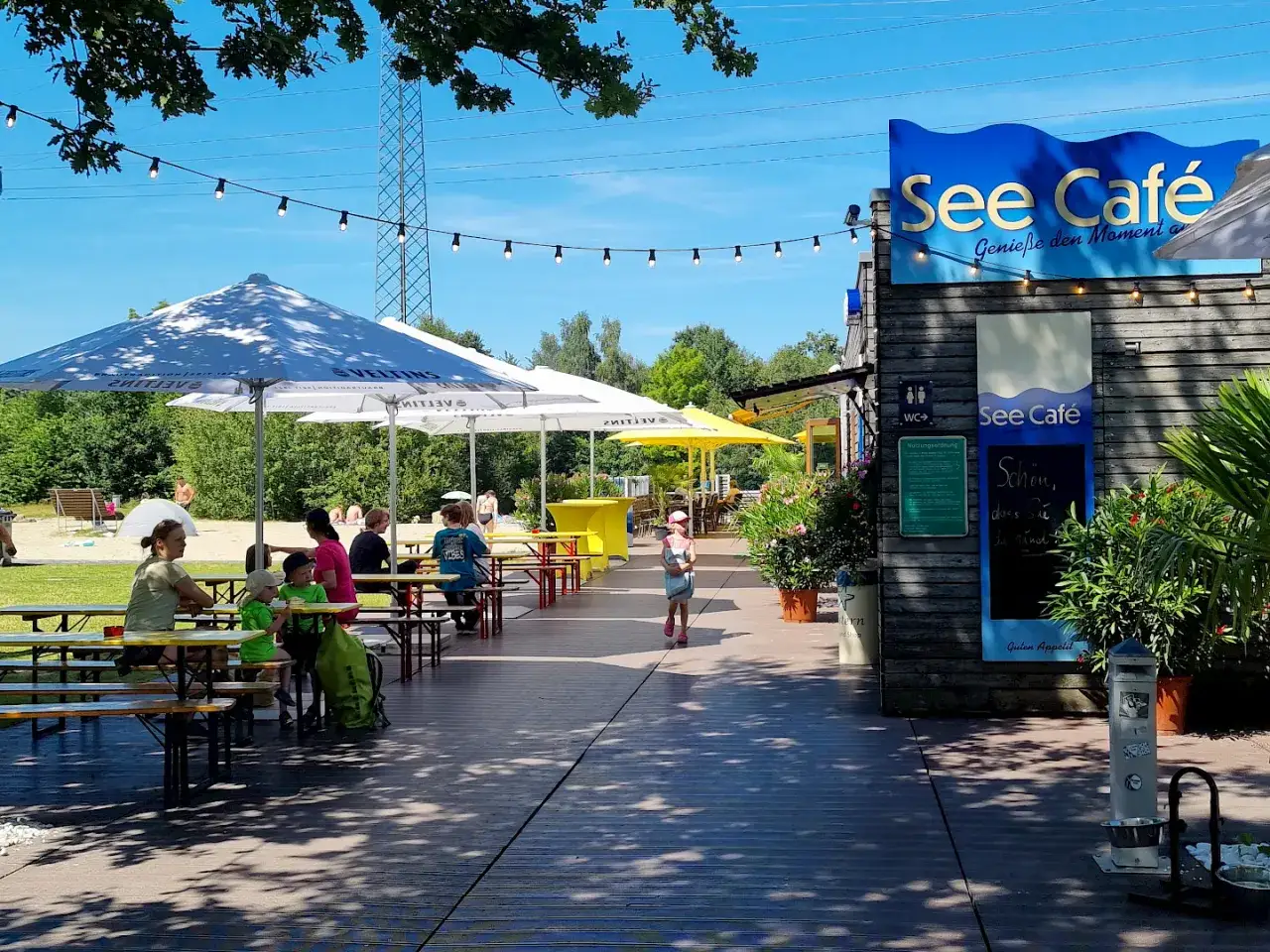 See Café mit Tischen und Gästen unter Sonnenschirmen am See.