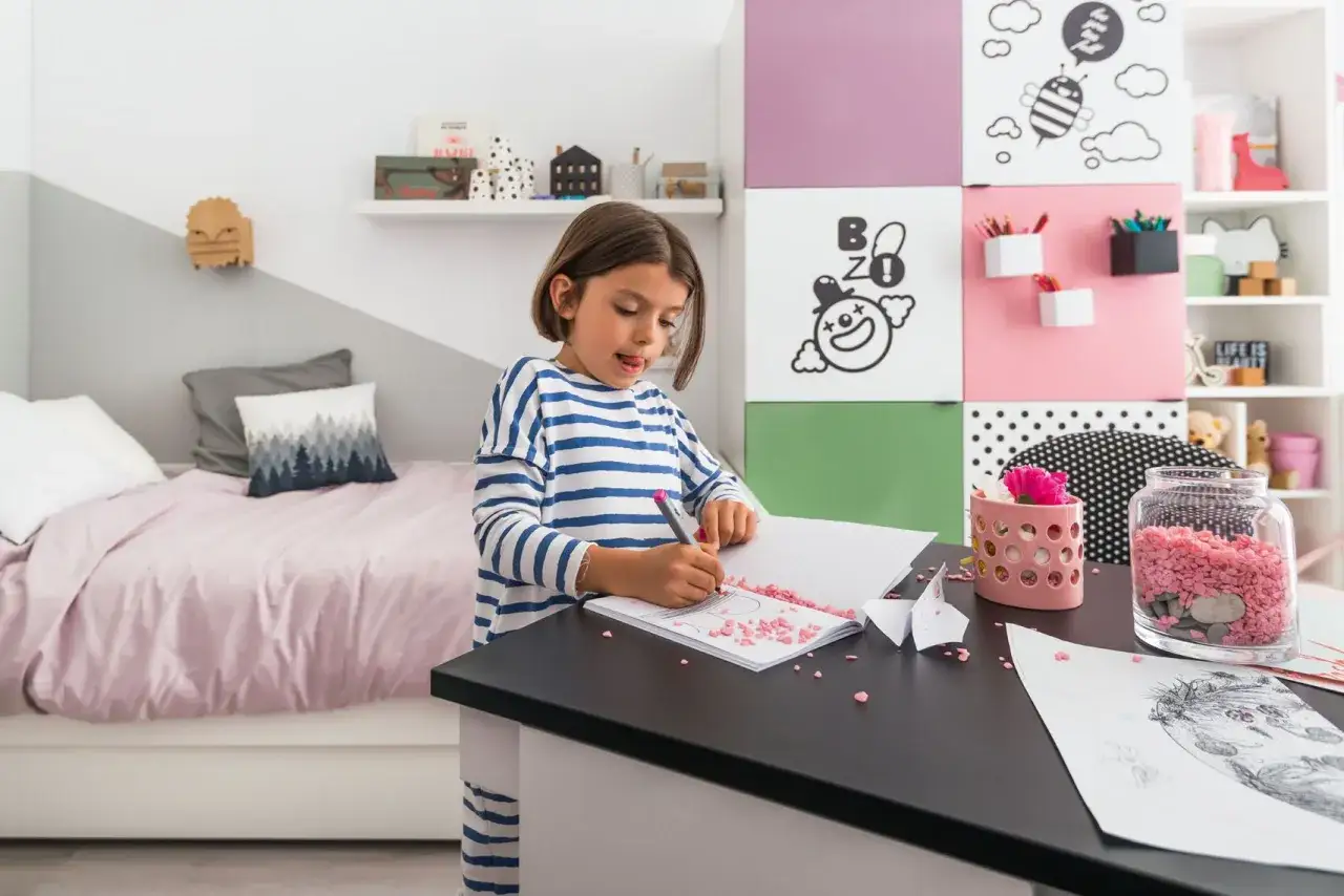 Dziewczynka rysuje w małym pokoju dziecięcym w bloku. Na ścianie kolorowe panele, na łóżku różowa pościel.