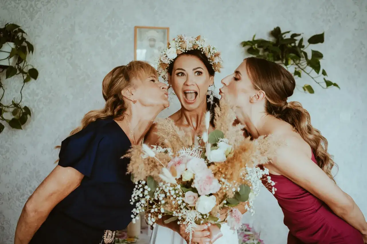 Panna młoda w wianku i dwie kobiety całujące ją w policzki, trzymają bukiet ślubny.