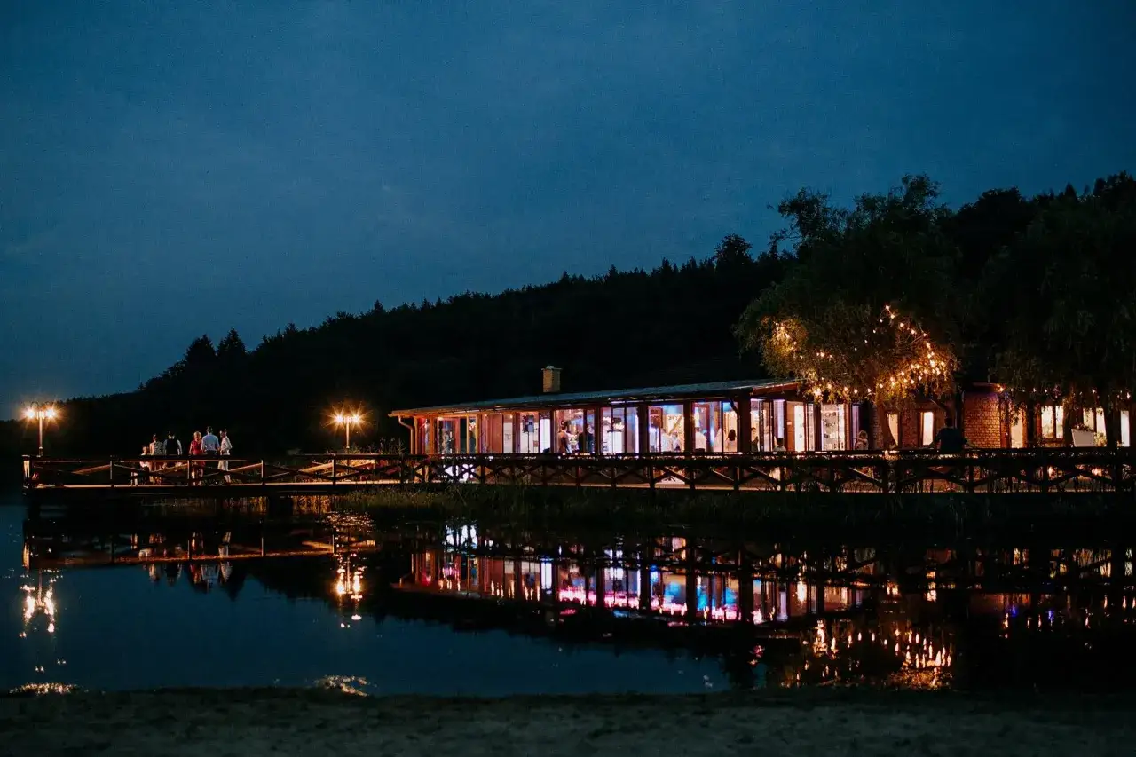 Wieczorny widok na budynek z przeszklonymi ścianami i oświetlonym molo nad jeziorem. Widać odbicia świateł w wodzie. To idealne miejsce na romantyczny wieczór w wodnym świecie Ostrzyce.
