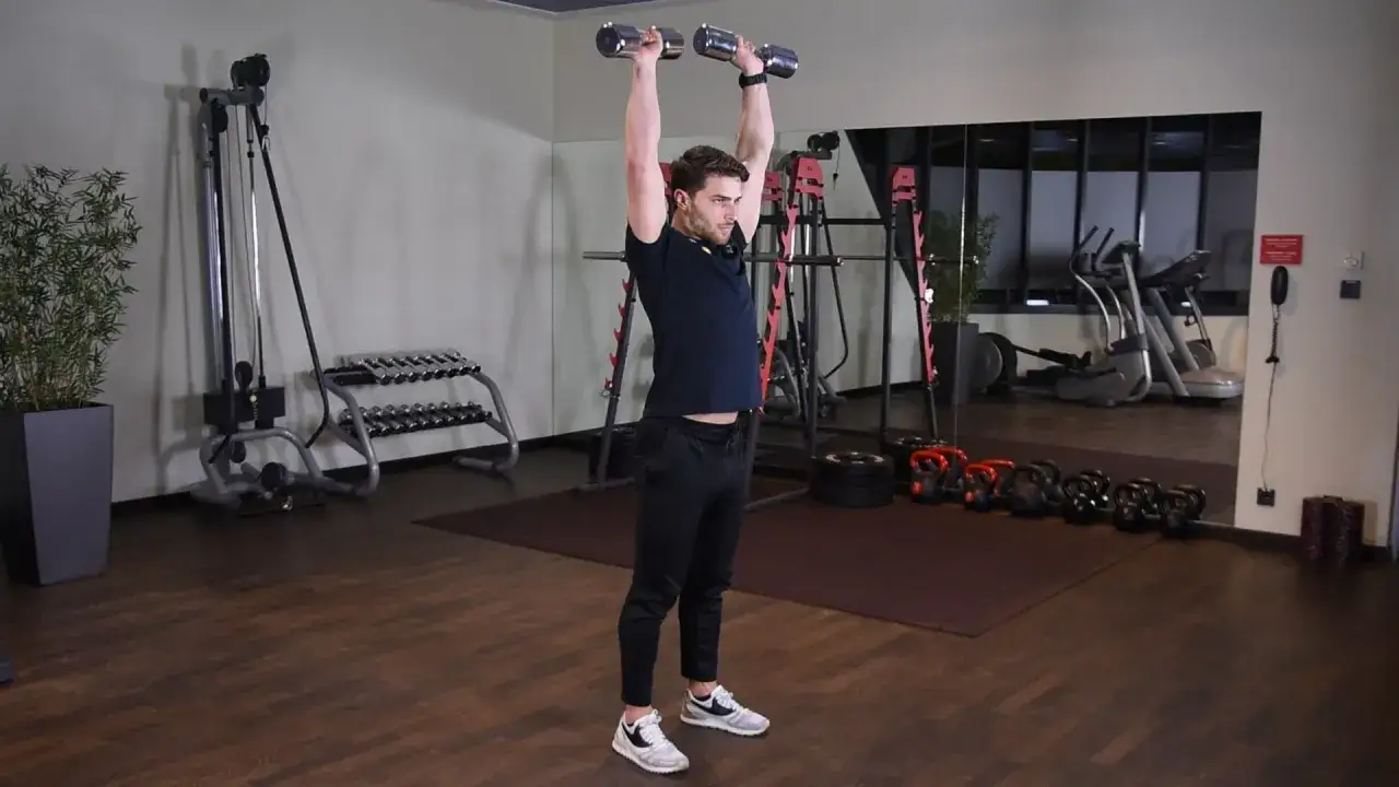 Wyciskanie hantli nad głowę angażuje mięśnie naramienne i triceps. Mężczyzna wykonuje ćwiczenie w siłowni.