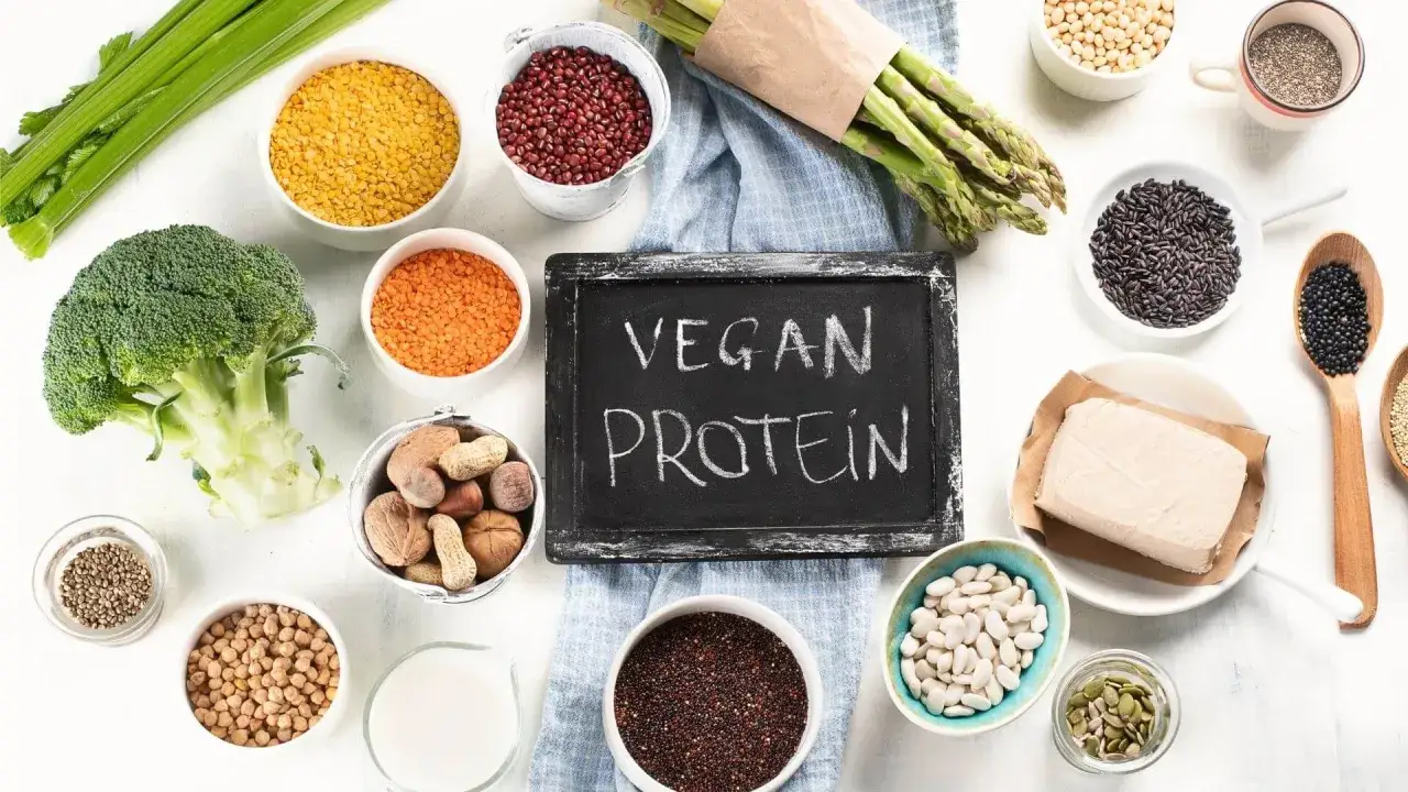 Tablica z napisem "VEGAN PROTEIN" otoczona miseczkami z roślinnymi źródłami białka: soczewicą, fasolą, orzechami, tofu, brokułem i szparagami.