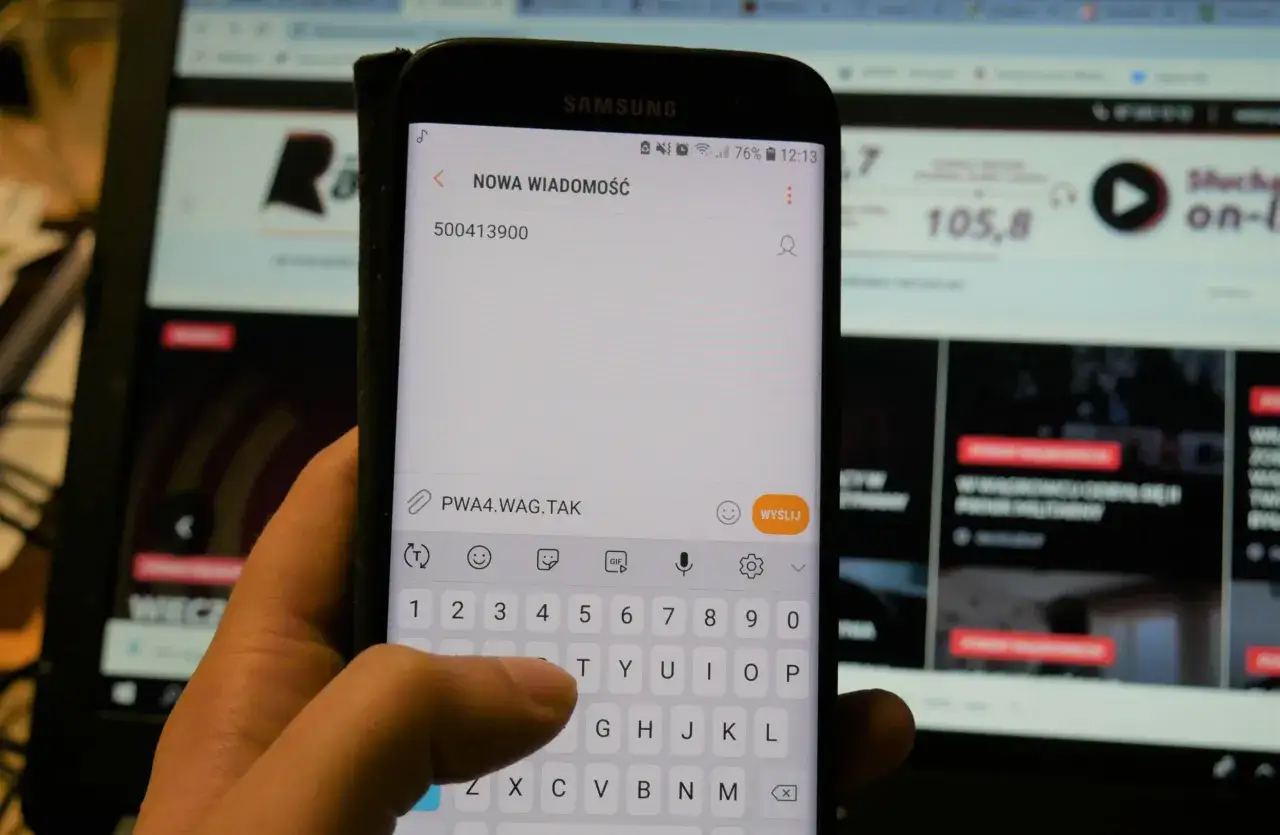 Dłoń pisze SMS na telefonie z numerem 500413900. W tle ekran komputera.