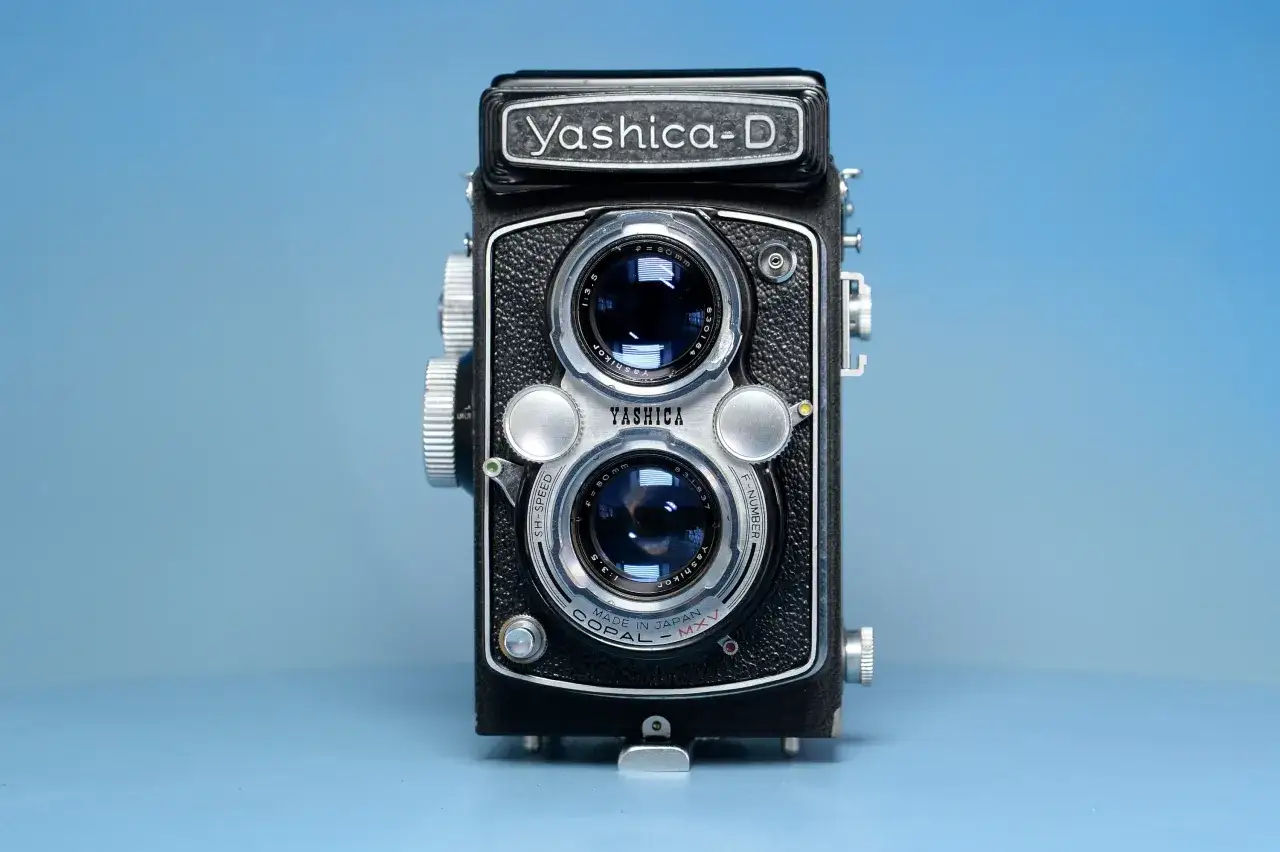 Czarno-srebrny aparat dwuobiektywowy Yashica-D na niebieskim tle.