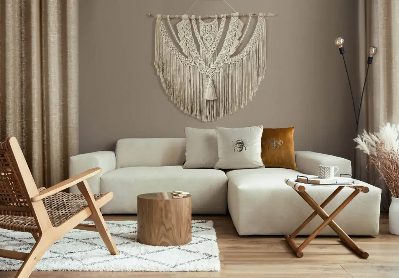 Makrama na ścianie to świetny pomysł, co powiesić na ścianie zamiast obrazu. Wnętrze z beżową sofą, drewnianym fotelem i dywanem.