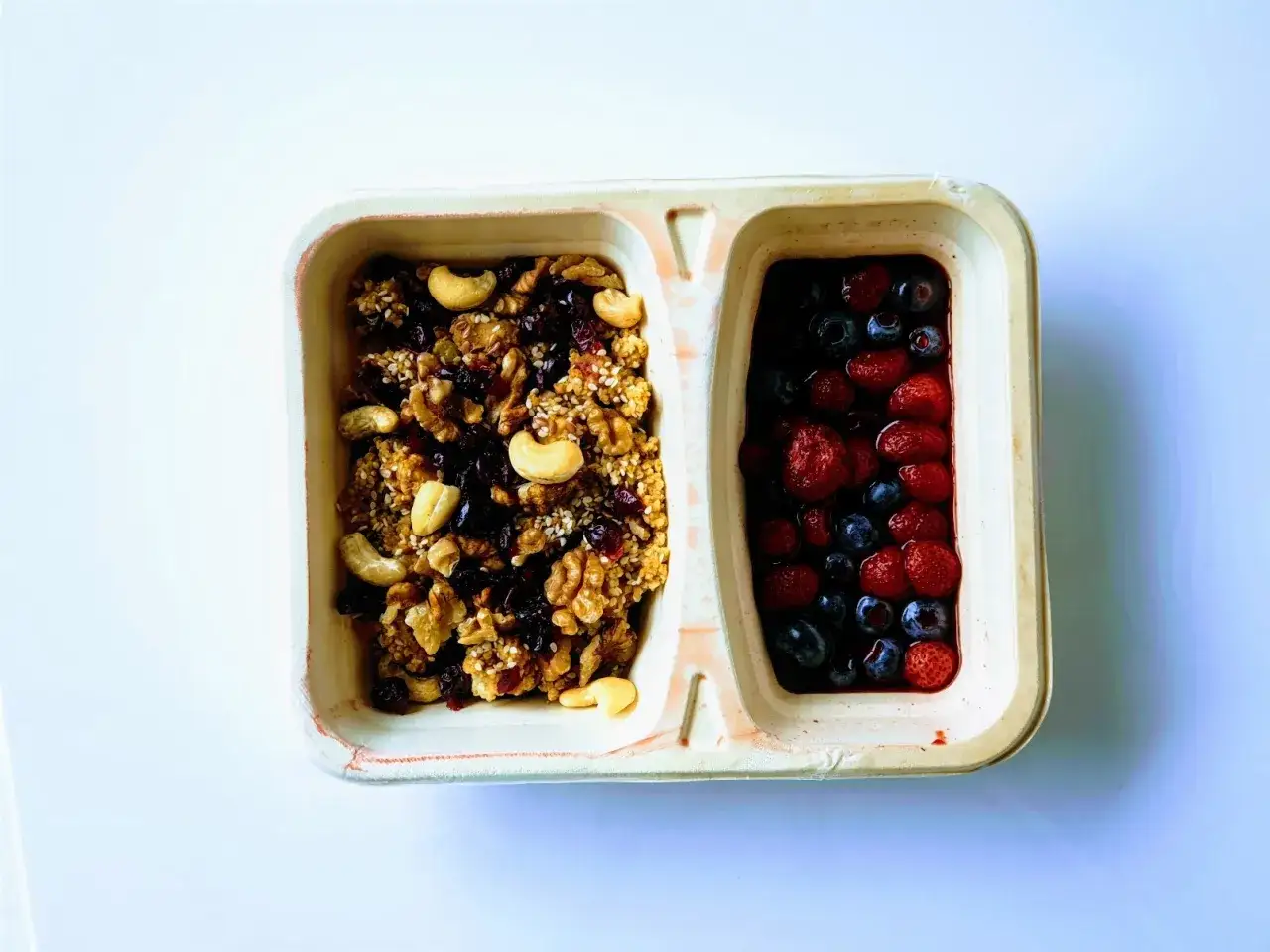 Dwie przegródki pojemnika: jedna z granolą i orzechami, druga z jagodami i truskawkami.