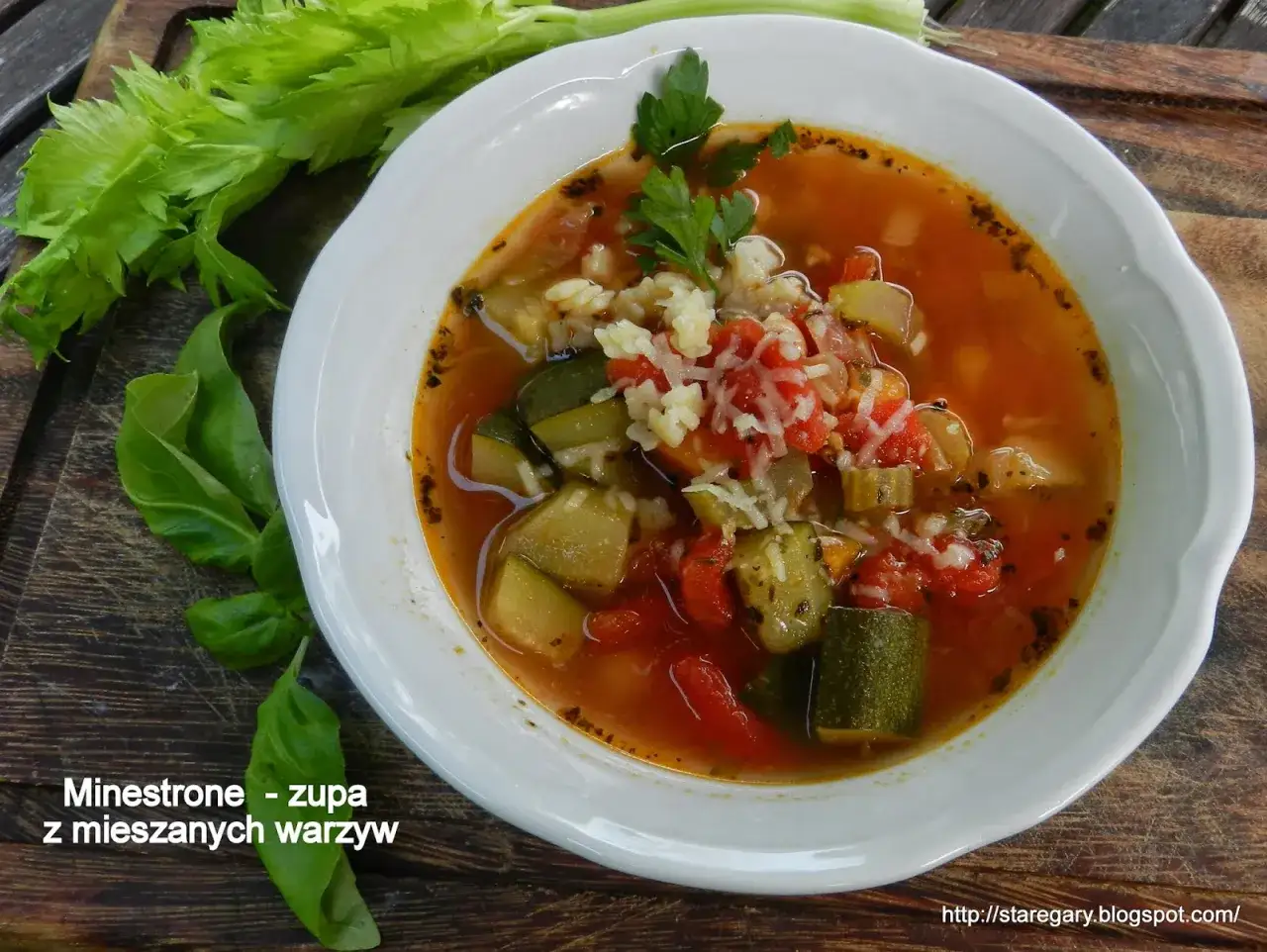 Miseczka zupy minestrone z warzywami i startym serem, obok łodygi selera i liście bazylii.
