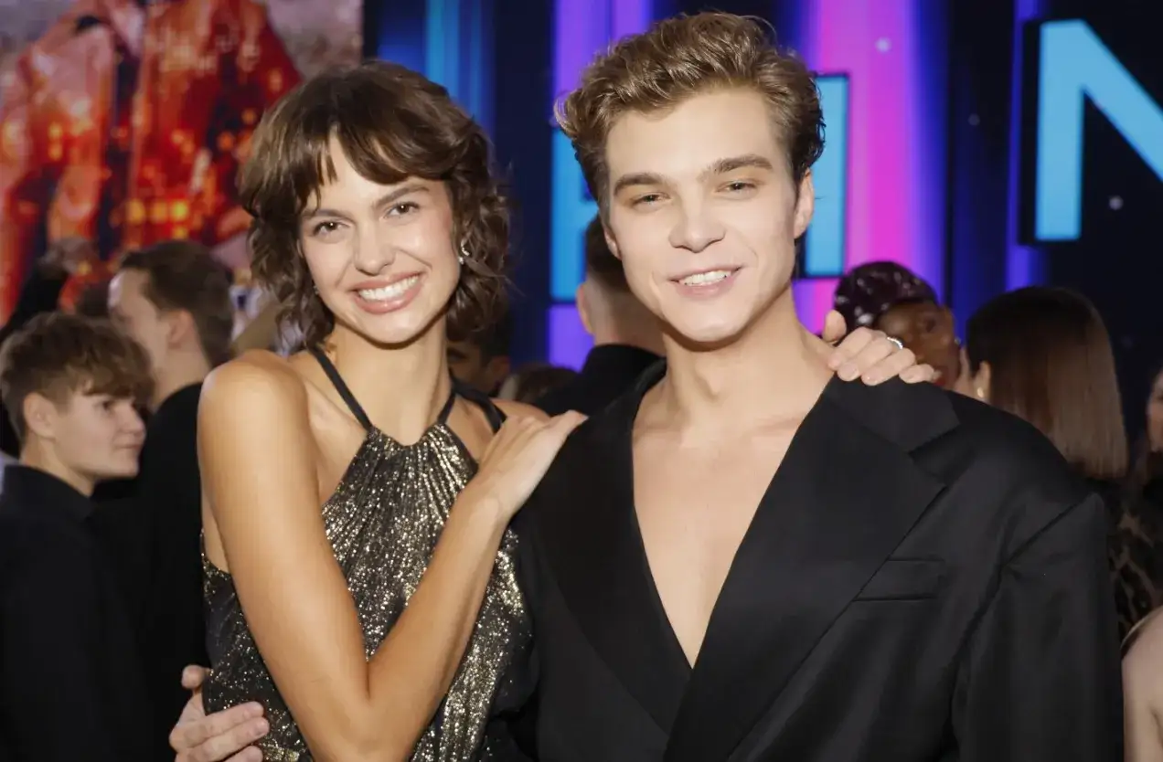 Uśmiechnięta para na evencie finał top model. Ona w błyszczącej sukience, on w eleganckim czarnym garniturze.