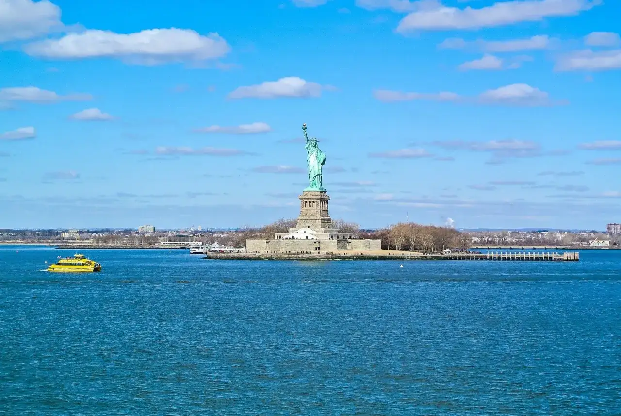 Statua Wolności na tle błękitnego nieba z chmurami. Obok żółta łódź. Zastanawiasz się, ile kosztuje lot do Stanów Zjednoczonych?