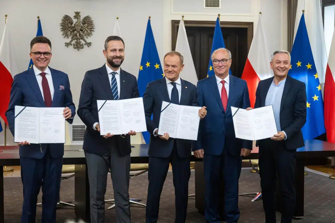 Pięciu mężczyzn w garniturach stoi przed flagami Polski i UE, trzymając dokumenty.