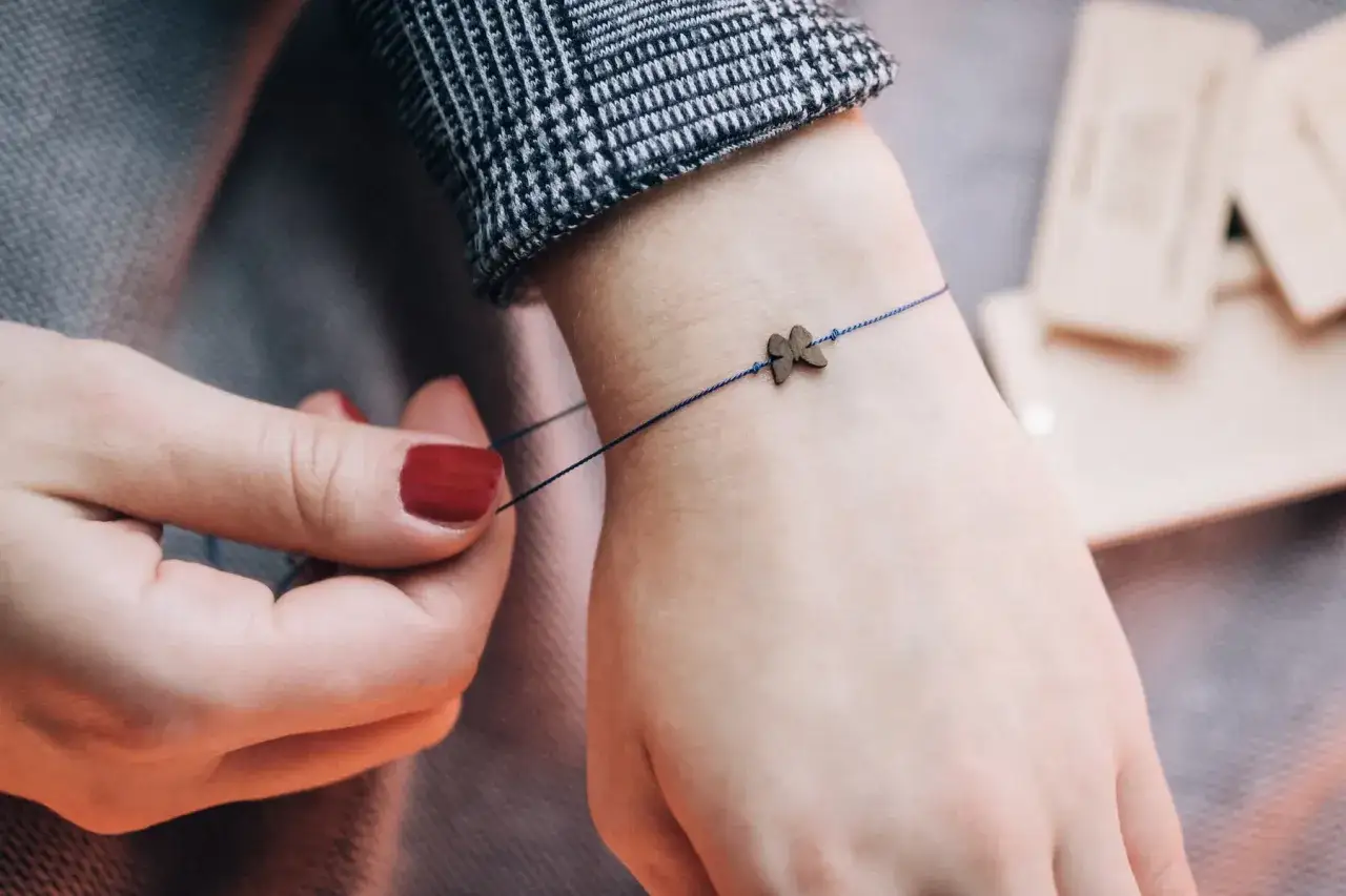 Dłoń z czerwoną emalią na paznokciach wiąże cienką, granatową nitkę z zawieszką w kształcie motyla.