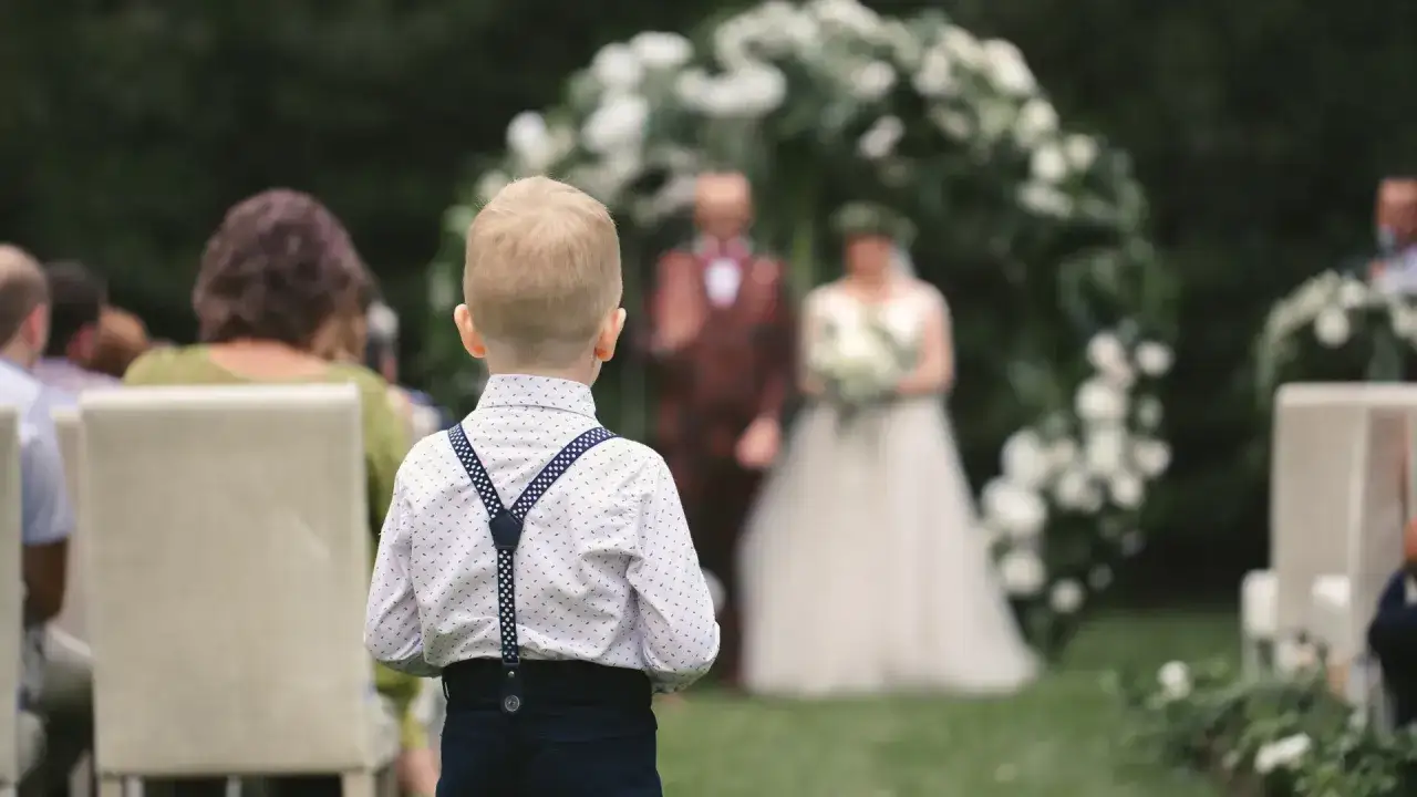 Chłopiec w białej koszuli z szelkami stoi tyłem do widza, na tle ślubu.