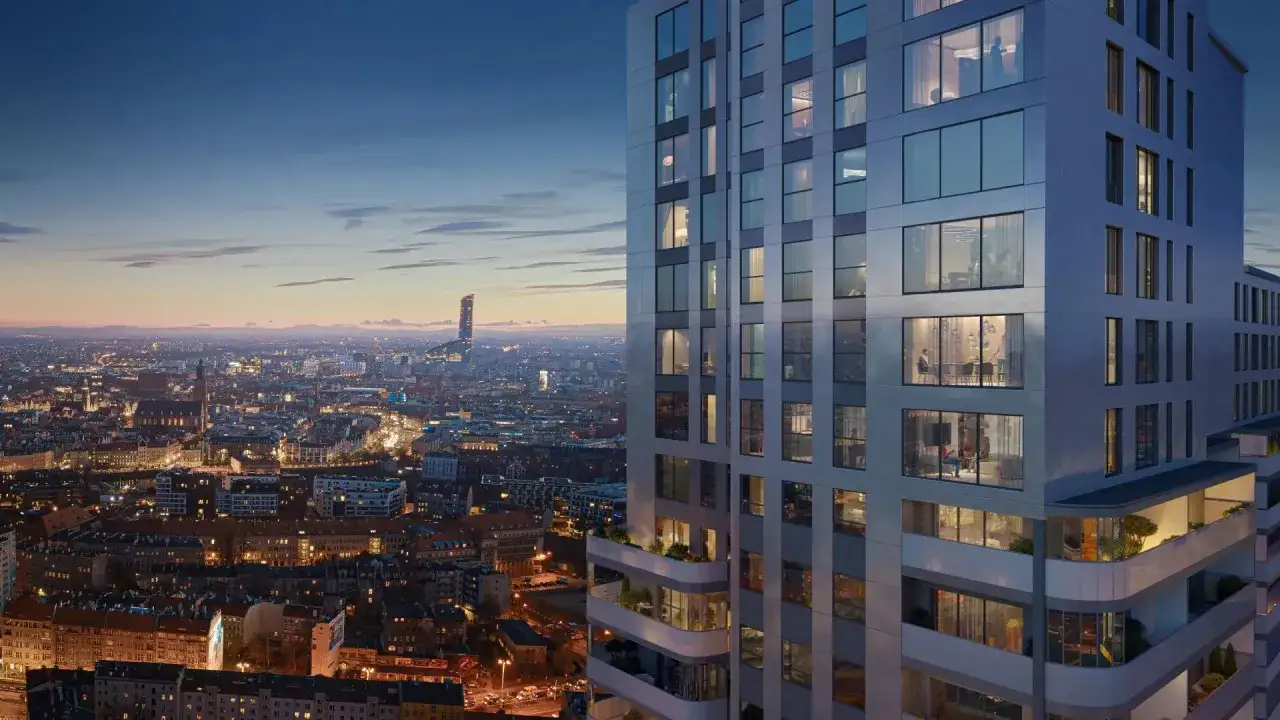 Nowoczesne osiedle apartamentowców w mieście nocą. Widać oświetlone okna, balkony z zielenią i panoramę miasta w tle.