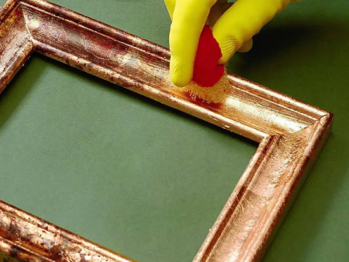 Dłoń w żółtej rękawiczce nakłada złoty lakier na ramę obrazu za pomocą gąbki.