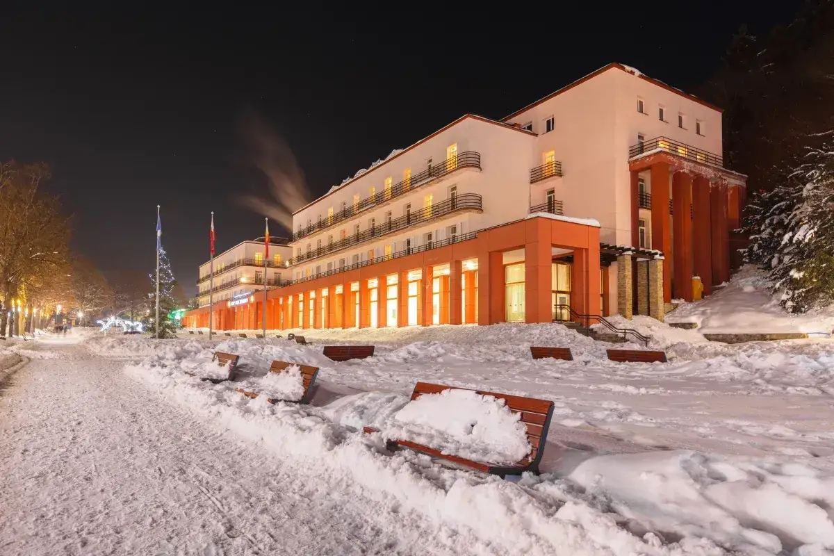 Sanatorium nocą, ośnieżone ławki i chodnik przed budynkiem.