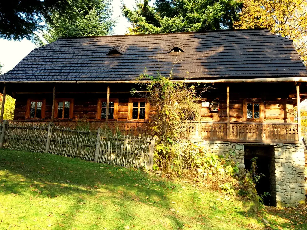 Drewniany dom z kamienną piwniczką i ozdobną werandą, otoczony zielenią i jesiennymi liśćmi.