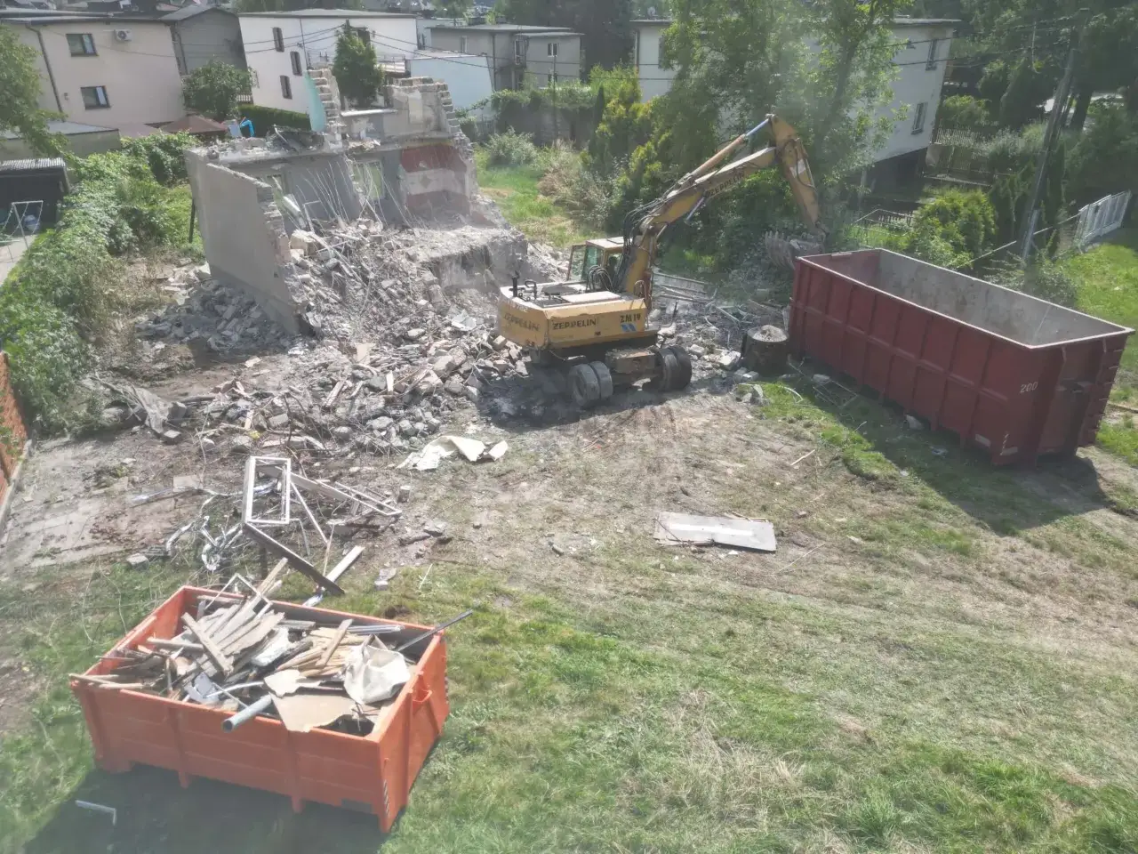 Koparka pracuje przy rozbiórce budynku. Gruz trafia do kontenerów. Oto jak zrobić opis rozbiórki.