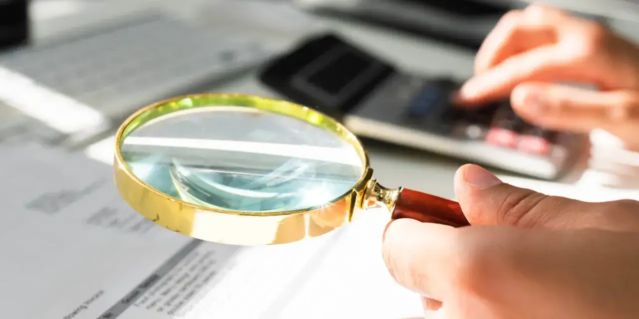 Dłoń trzymająca lupę nad dokumentami, w tle kalkulator i klawiatura.