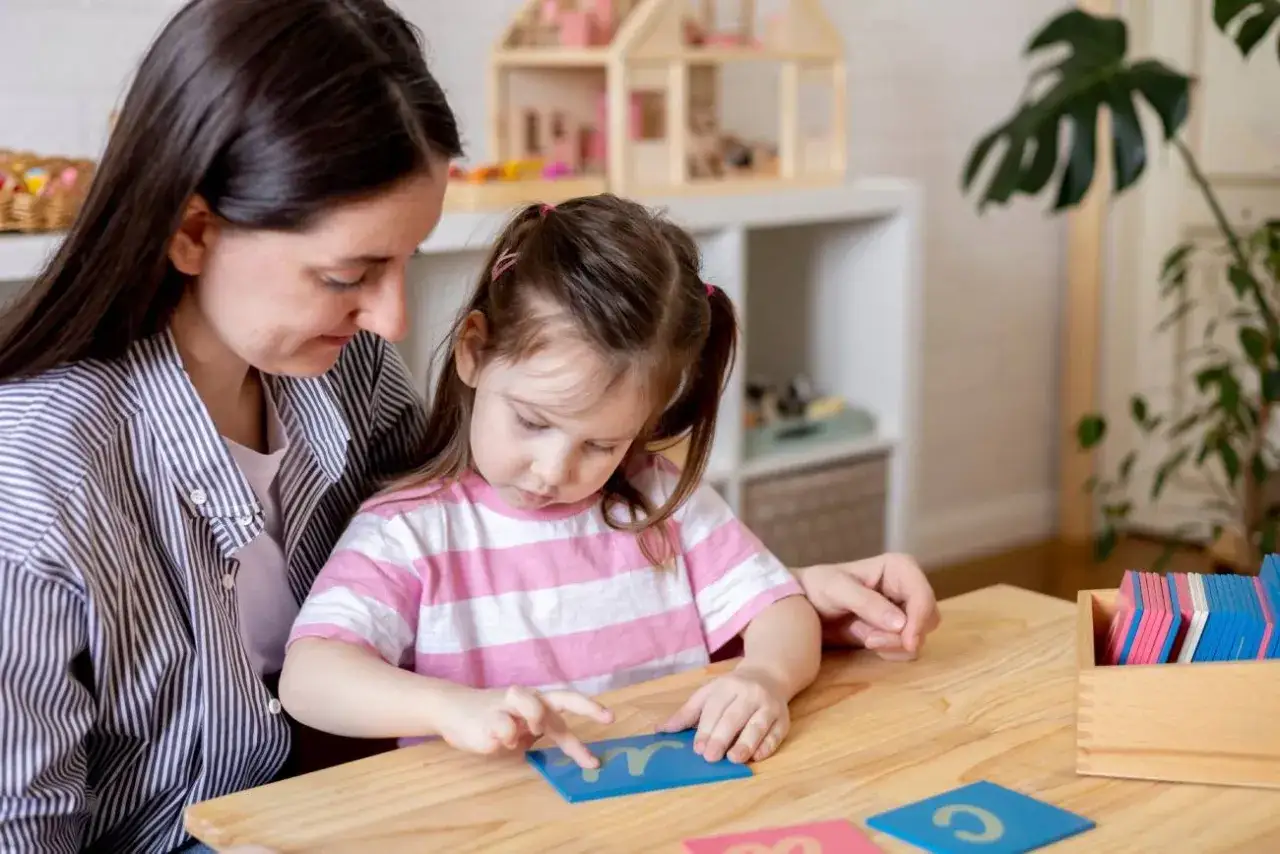 Mama uczy córkę liter, przygotowując ją do przedszkola. Dziecko z uwagą śledzi paluszkiem literkę na niebieskiej karcie.
