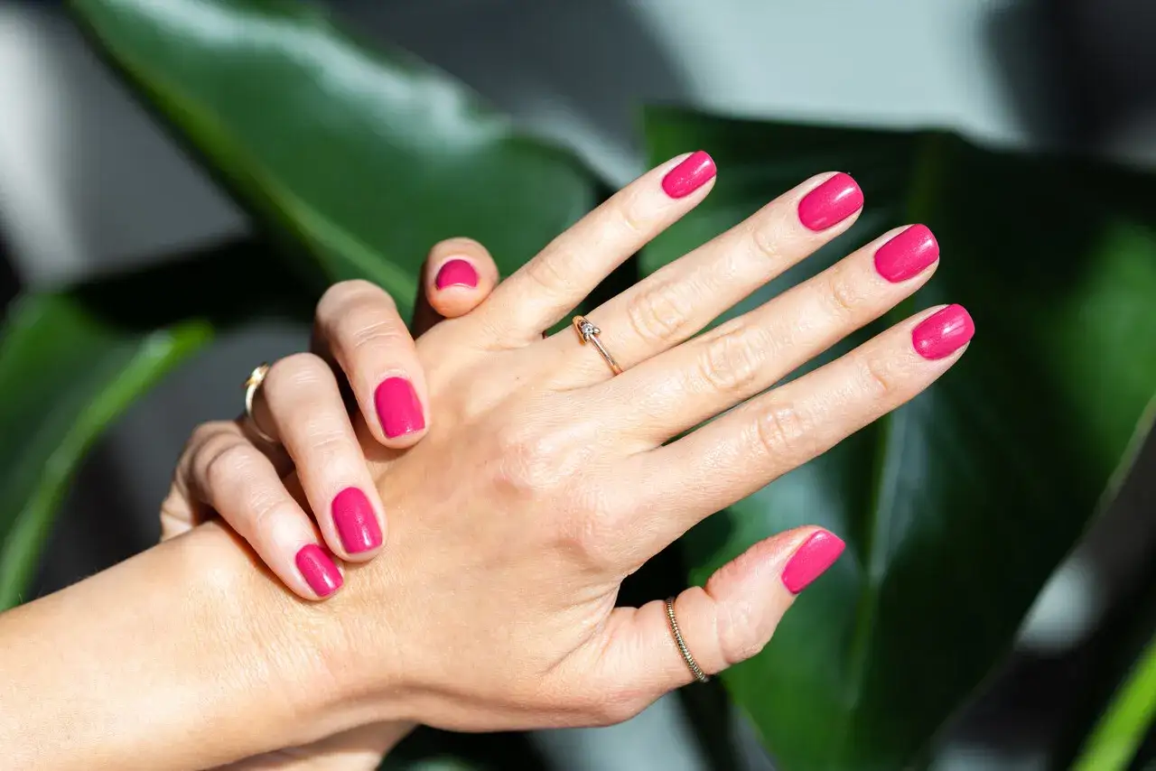 Dłonie z idealnie pomalowanymi na fuksję paznokciami. W tle bujne, zielone liście. Idealny wybór co na paznokcie dla odważnych.