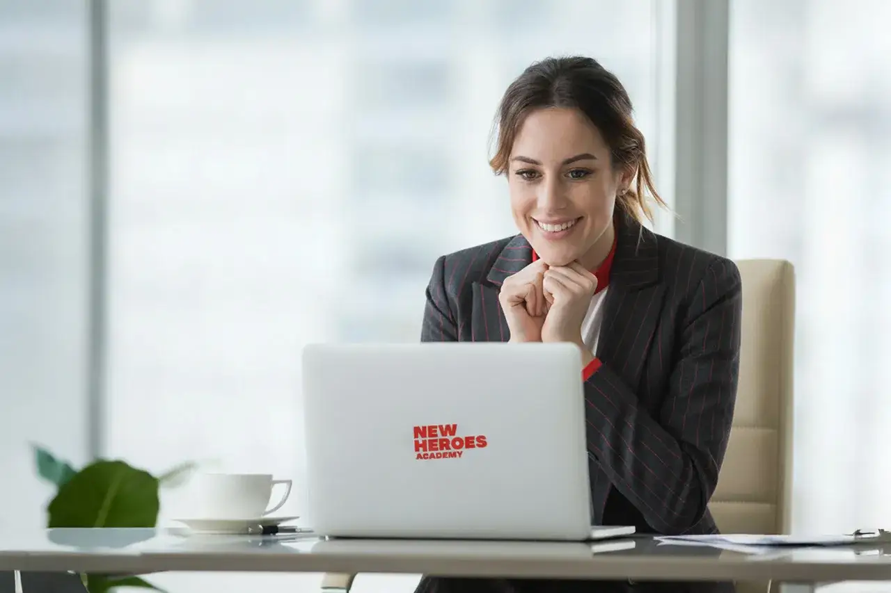 Uśmiechnięta kobieta w garniturze patrzy na laptopa z logo "New Heroes Academy".