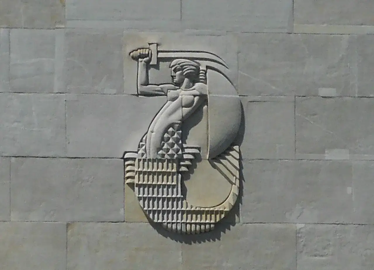 Warszawska Syrenka w płaskorzeźbie na kamiennej ścianie, trzymająca miecz i tarczę.