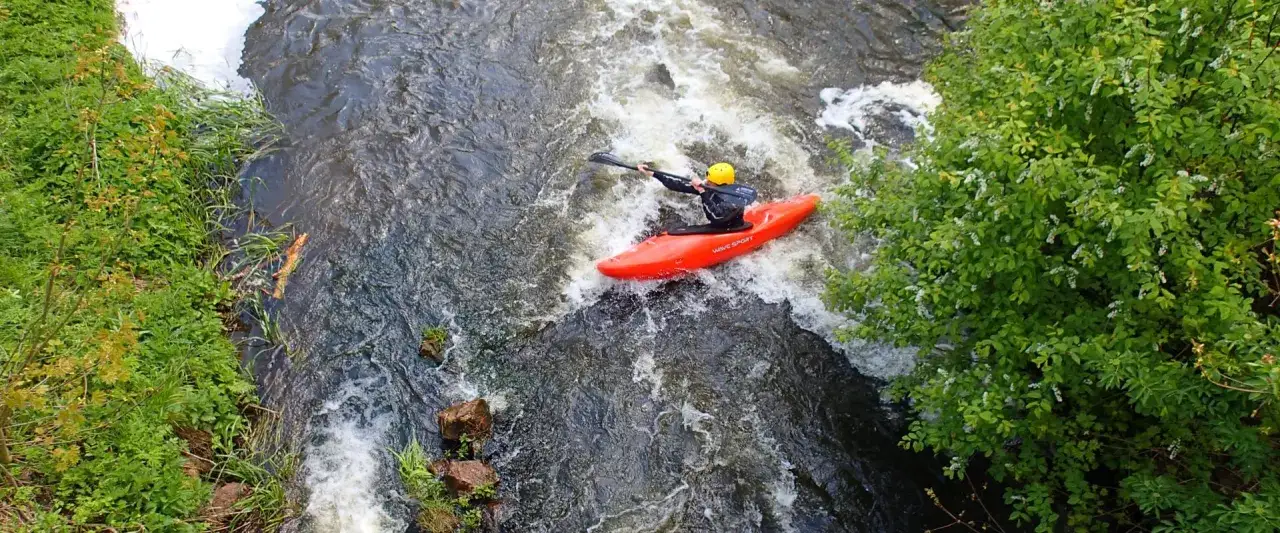 Kajakarz w żółtym kasku płynie czerwonym kajakiem po rwącym nurcie rzeki.