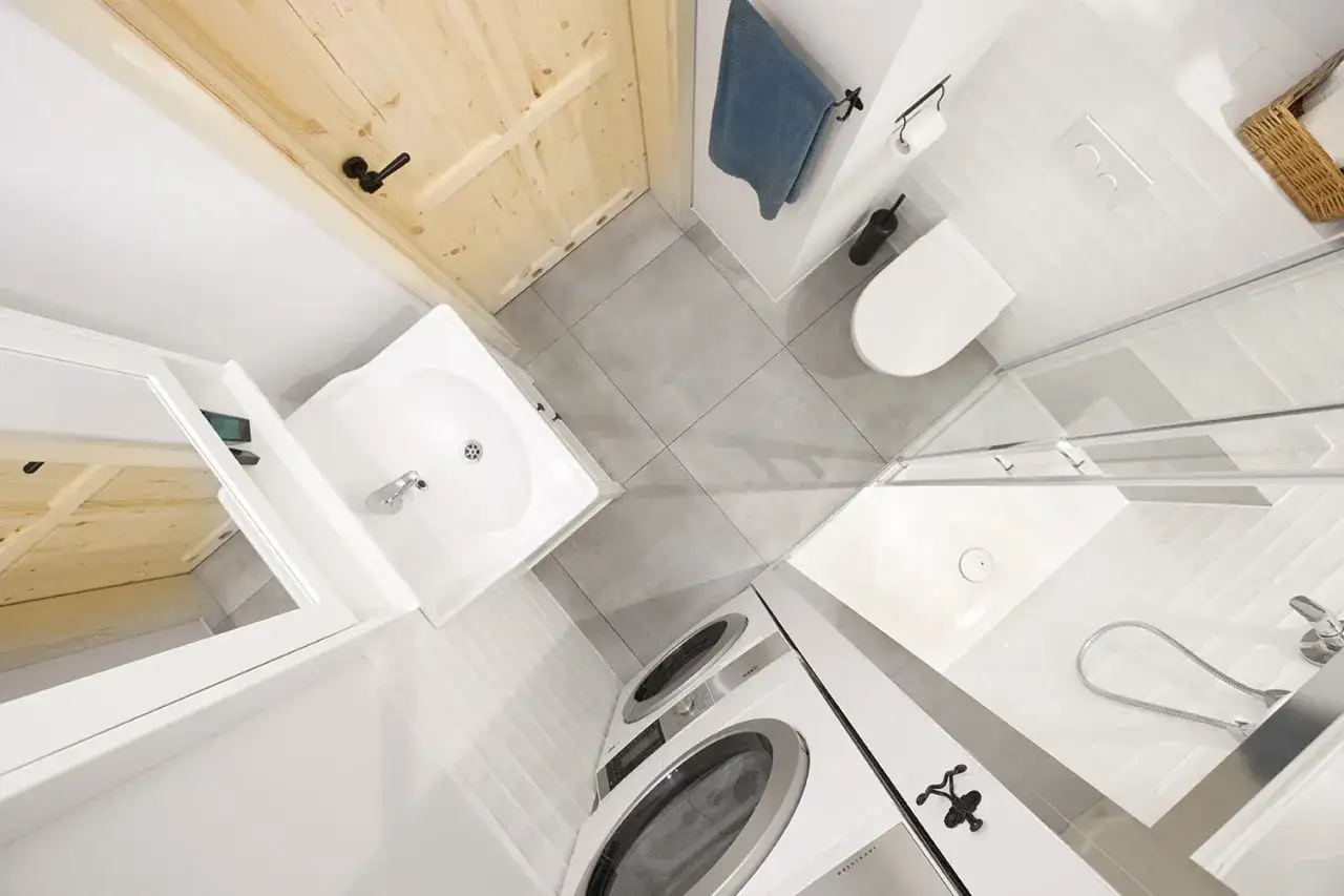 Praktyczna aranżacja małej łazienki z prysznicem i pralką w bloku. Drewniane drzwi dodają ciepła.