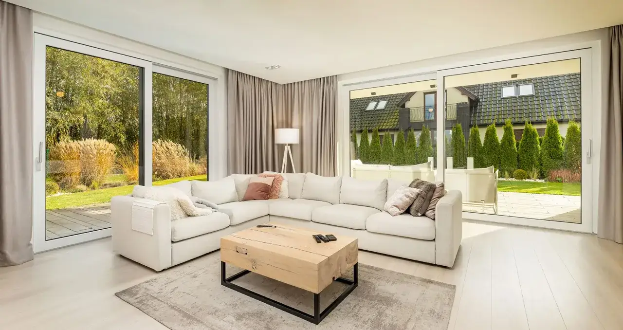 Nowoczesna aranżacja drzwi balkonowych z wygodną sofą i drewnianym stolikiem. Widok na ogród i domy.