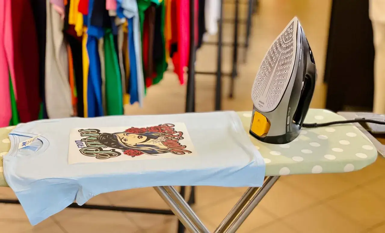 Jak zrobic naprasowanke? Na desce do prasowania leży jasnoniebieska koszulka z nadrukiem, obok żelazko.