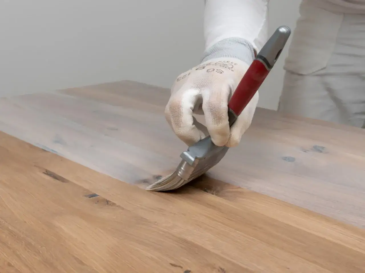 Ręka w rękawiczce nakłada biały preparat na drewniany blat, pokazując, jak usunąć wosk z drewna.