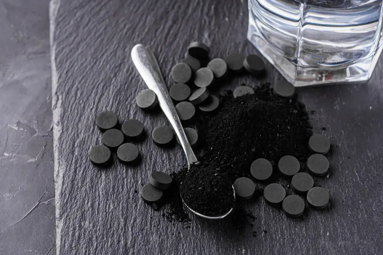 Węgiel aktywny w proszku i tabletkach na ciemnym tle, obok łyżeczki i szklanki wody. Pokazuje, jak działa węgiel aktywny.