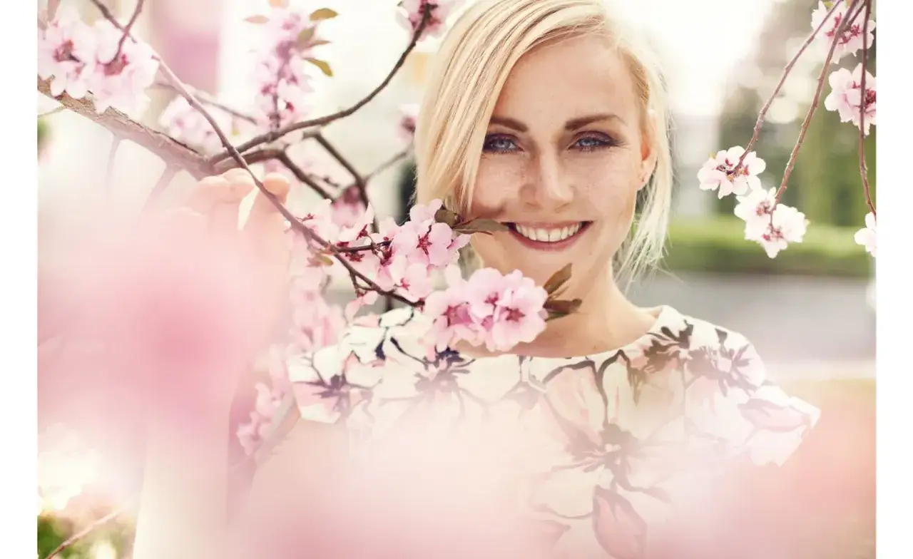 Uśmiechnięta blondynka wśród kwitnących gałązek wiśni. Jej twarz, ozdobiona piegami, promienieje radością, idealnie oddając, jak wygląda pani wiosna.