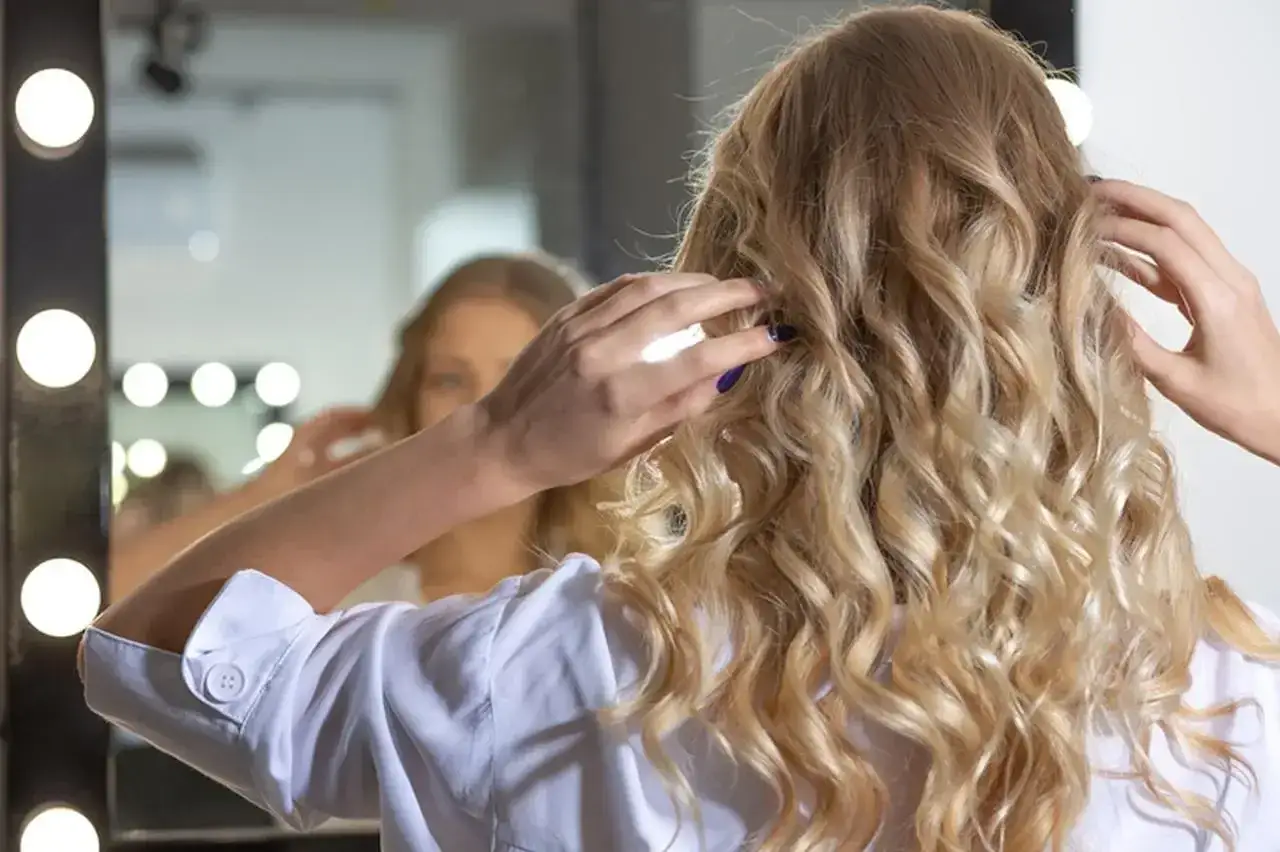 Blondynka poprawia nowoczesne fale na włosach przed lustrem z lampkami.