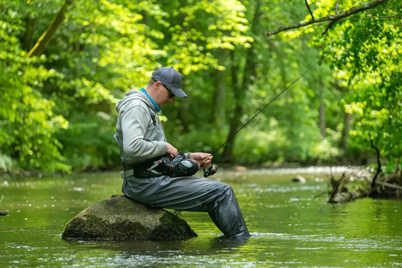 Wędkarz siedzi na kamieniu w rzece, łowiąc ryby. Pamiętaj, że ile kosztuje mandat za brak karty wędkarskiej, by uniknąć problemów.