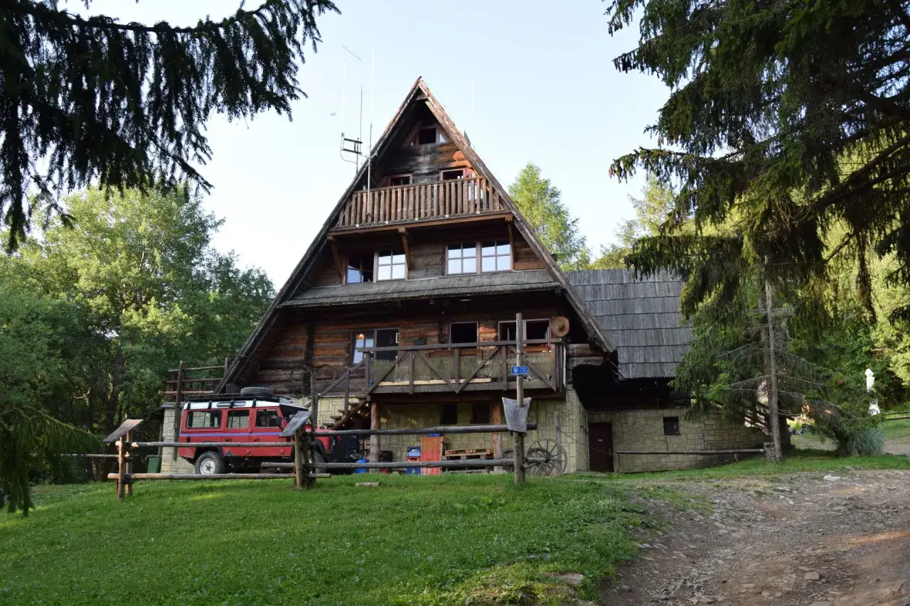 Drewniana bacówka pod Małą Rawką, z czerwonym Land Roverem zaparkowanym obok. Otoczona zielenią i drzewami.