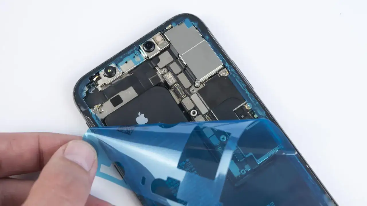 Dłoń odkleja niebieską folię od wnętrza iPhone'a XR z widoczną baterią.