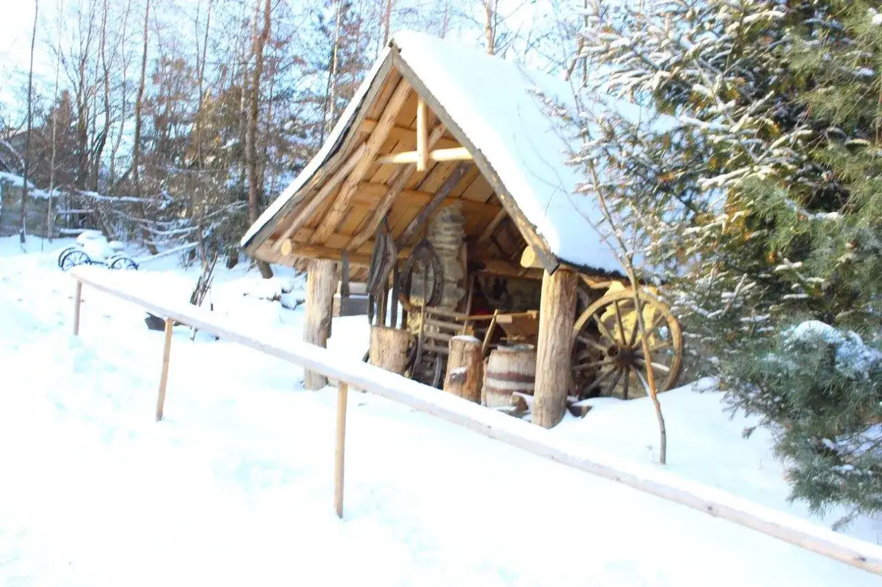 Zimowa sceneria wokół bacówki u harnasia. Drewniana chata kryta śniegiem, w środku narzędzia i beczki.