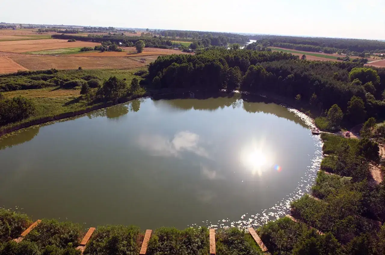 Widok z lotu ptaka na jezioro otoczone lasem i polami, z widocznymi pomostami.