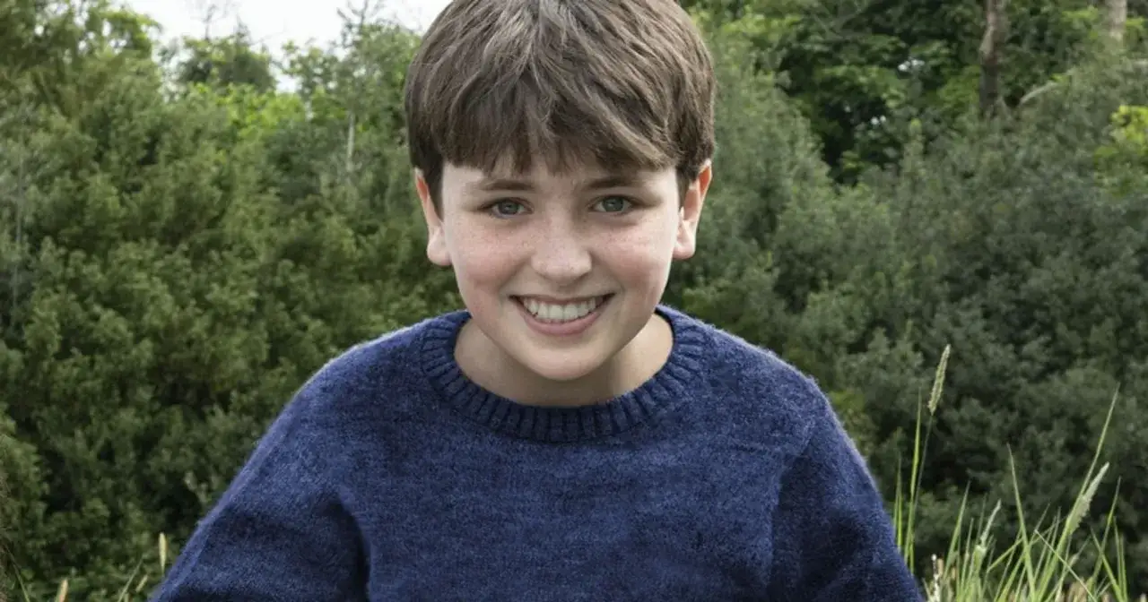 Uśmiechnięty chłopiec w granatowym swetrze, jeden z młodych aktorów Harry'ego Pottera, na tle zielonego lasu.